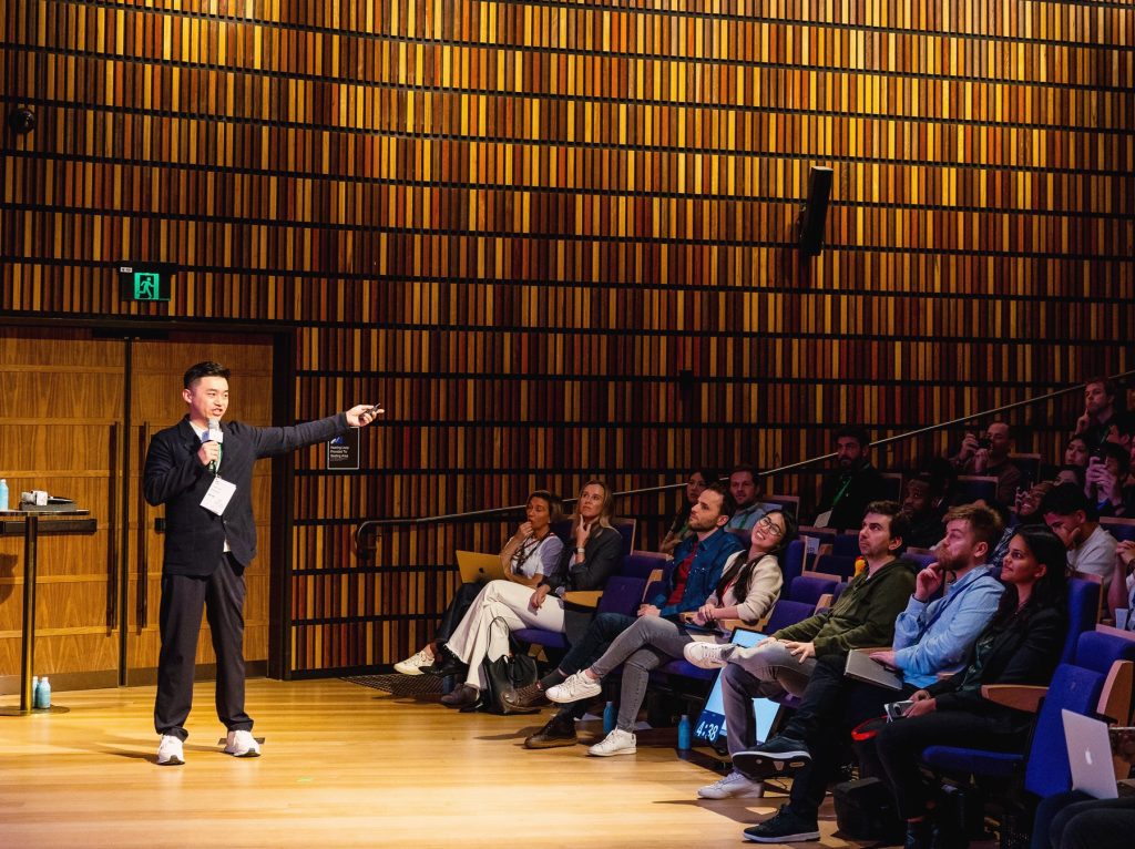 A speaker pointing to a presentation in front of a seated audience in a wood-panelled auditorium
