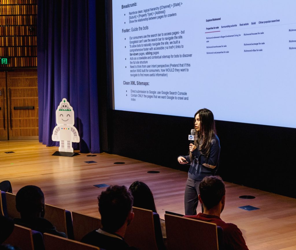 An SEO speaker with a microphone presenting on a conference stage, a slide about site structure behind her and the audience in the foreground